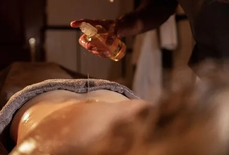 Spa Treatment with Oil at GRANDSPA Zell am See A person pours oil onto the back of a lying guest during a spa treatment in a warm spa room.